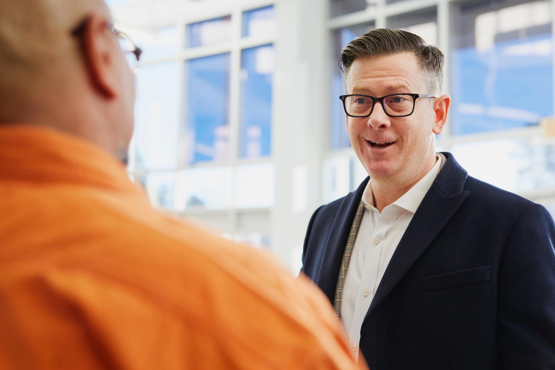 A man in a suit and glasses is talking to another man in an orange shirt.